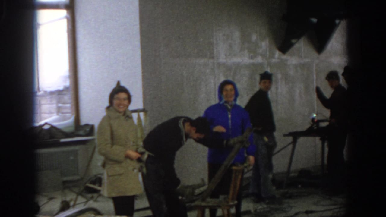 A smiling woman stands near a man working as other adults remain in the unfinished room.