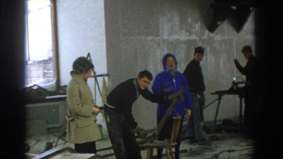 Munich 1963 home-movie frame with a man looking toward the camera, a woman in blue nearby, and tools in an unfinished interior.