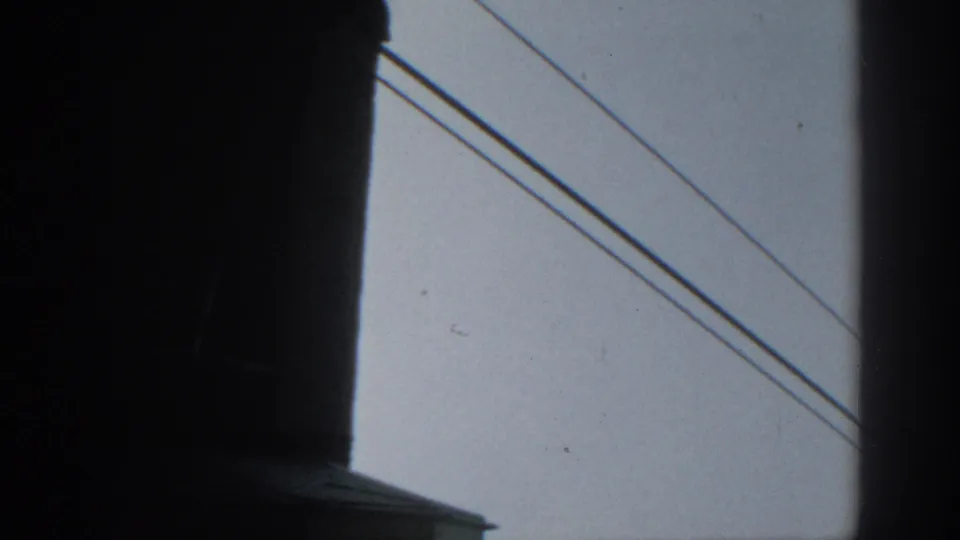 1963 Munich home-movie frame showing silhouetted foreground structure and power lines crossing a gray sky.