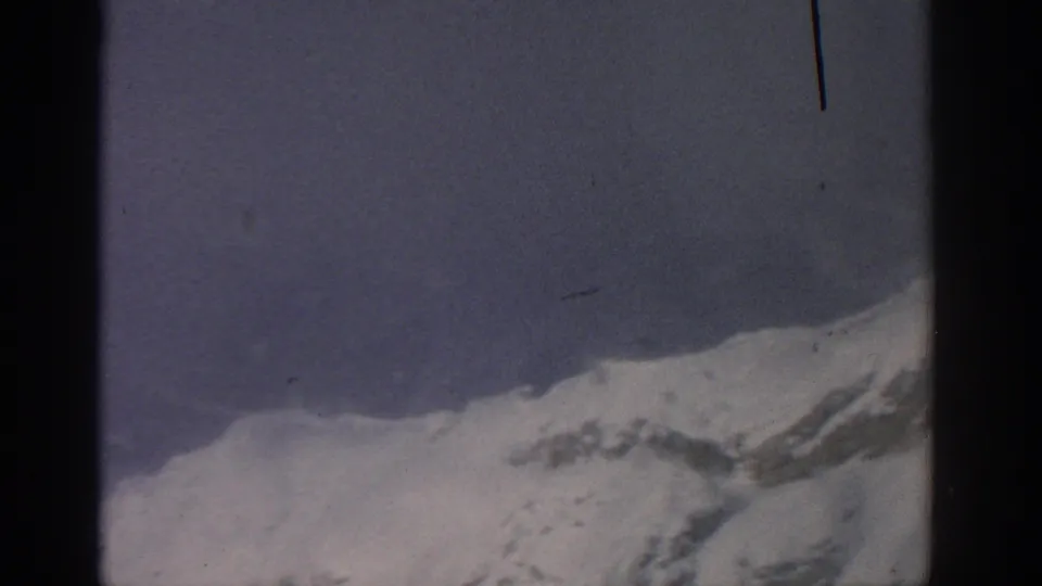 1963 8mm aircraft-view frame showing a snow-covered mountain ridge below pale sky, divided by dark film gate edges.