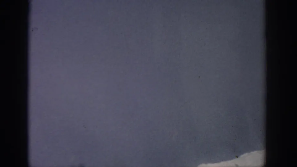 Aircraft-window view in the 1963 home movie, with pale blue sky and a small snowy mountain edge at lower right.