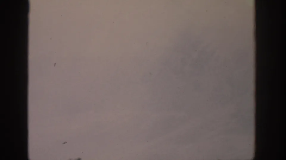 1963 8mm aerial view with pale sky above a soft, snow-covered mountain surface and visible film-frame borders.