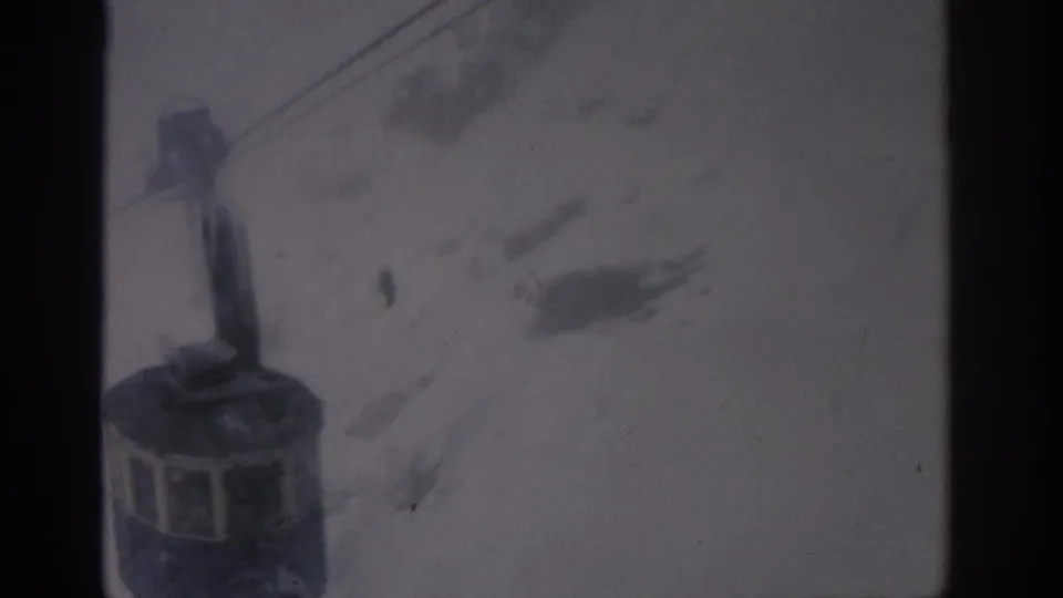 1963 Munich-labeled 8mm footage shows a lift cabin or chair near overhead cables with snowy terrain behind it.