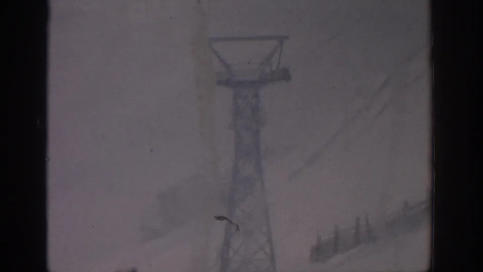 1963 Munich-labeled 8mm frame showing a chairlift support tower, overhead cables, and snowy terrain in soft focus.