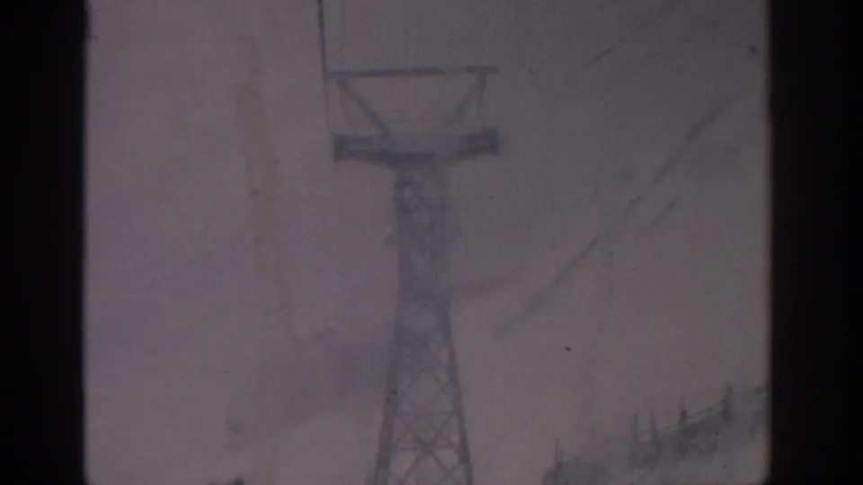 1963 Munich home-movie frame showing a metal cable car or ski-lift tower fading into a gray snowstorm.