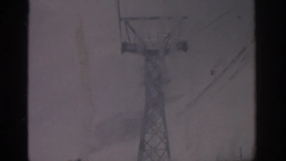 A snowy 1963 Munich frame shows the upper section of a metal lift tower with cables against a muted winter sky.