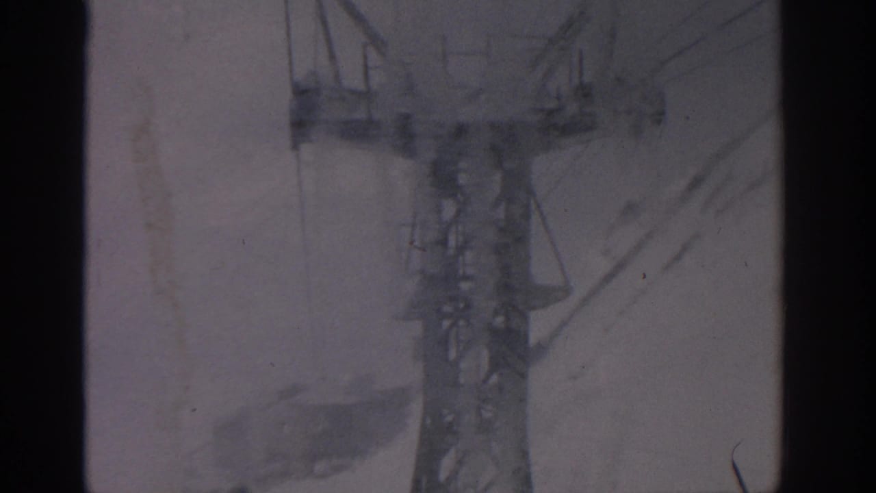 1963 8mm cable-car view near Munich, with a steel support tower and cables seen through dark window framing over snow.