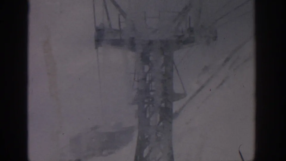 1963 8mm cable-car view near Munich, with a steel support tower and cables seen through dark window framing over snow.