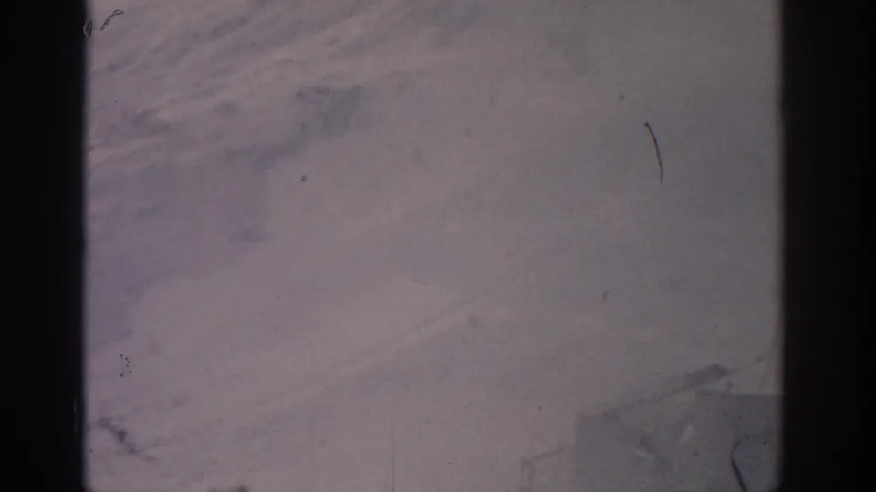 Snow-covered terrain fills the view from inside the moving cable car, with dark window borders and soft 8mm grain.