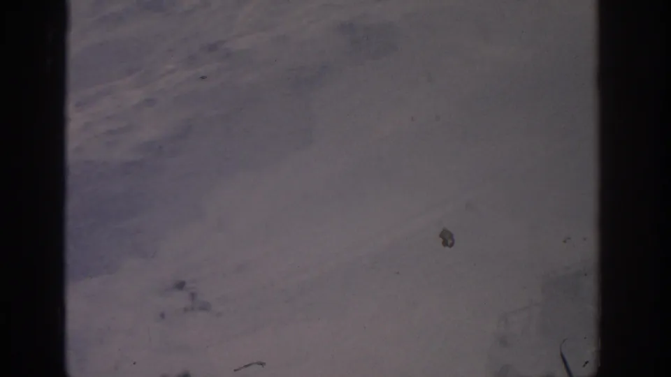 1963 8mm frame showing a pale, open landscape with mottled darker patches and visible film gate edges.