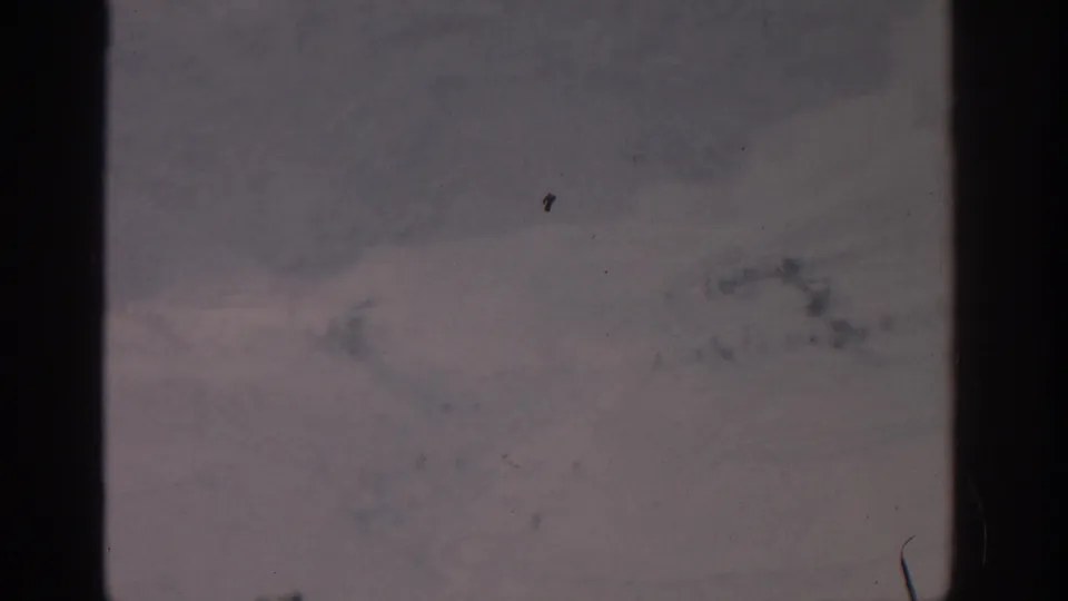 1963 Munich 8mm frame with a muted snowy expanse, indistinct terrain marks, dust, scratches, and dark vertical frame edges.