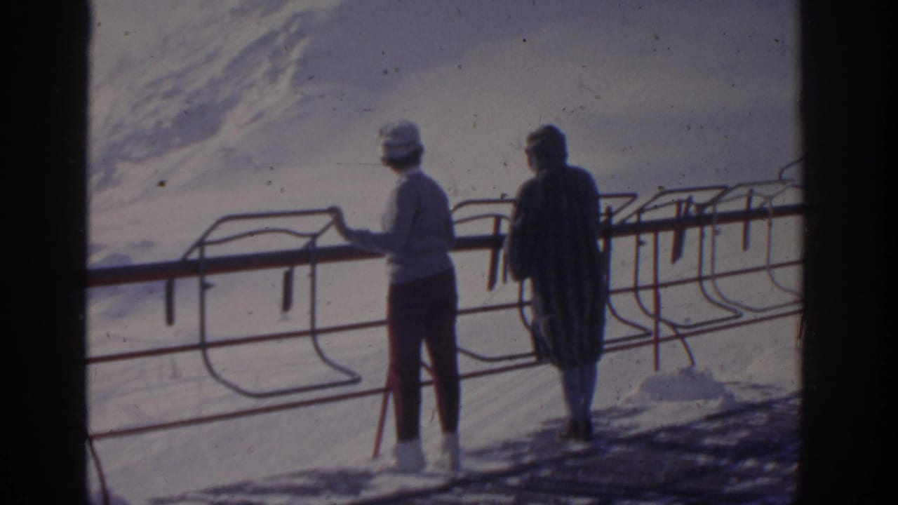 1963 home-movie frame of two women near a ski-area railing with snow-covered mountains behind them.