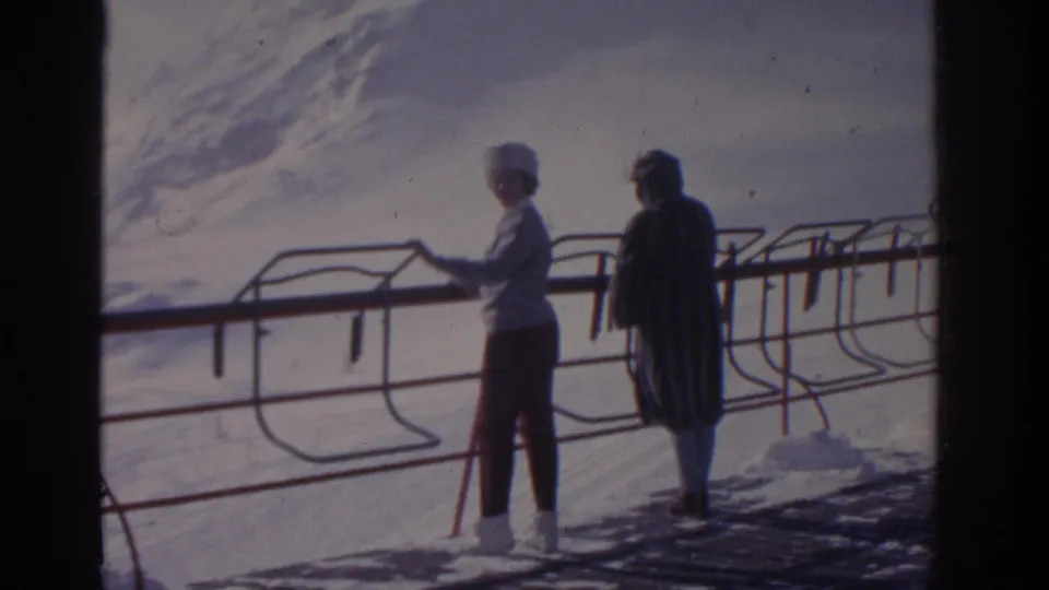 A woman in a white hat faces the snowy landscape while her companion stands near a metal railing in 1963 Germany footage.
