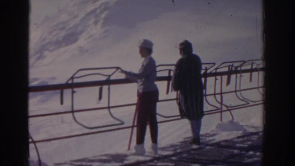 The white-hatted woman gestures toward the snow while standing beside a companion at a mountain railing in 1963 home-movie footage.
