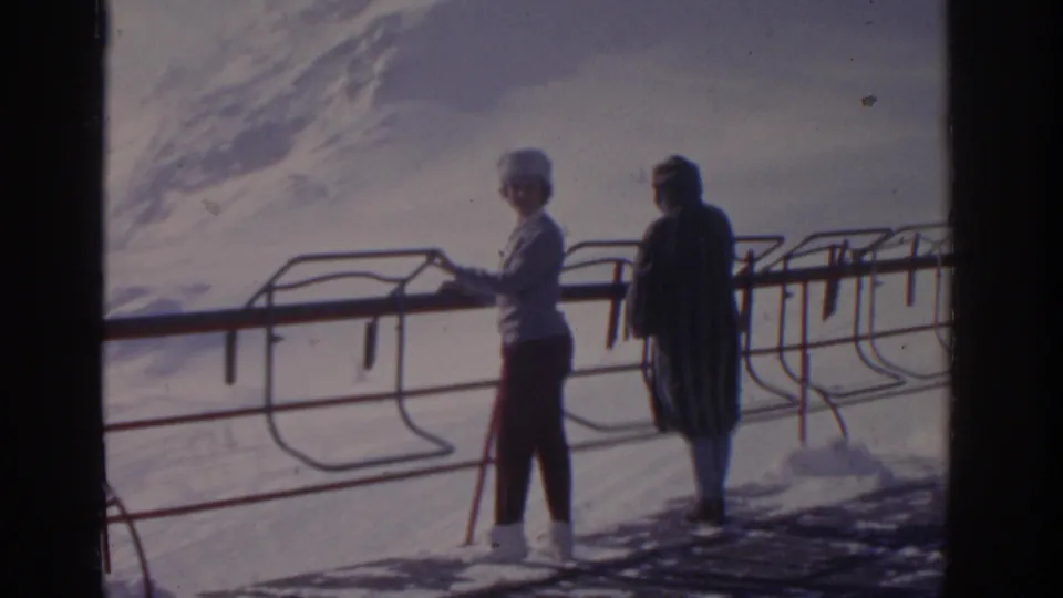 Two women remain beside the metal railing on a snowy mountain slope, framed by 8mm film edges and soft winter light.