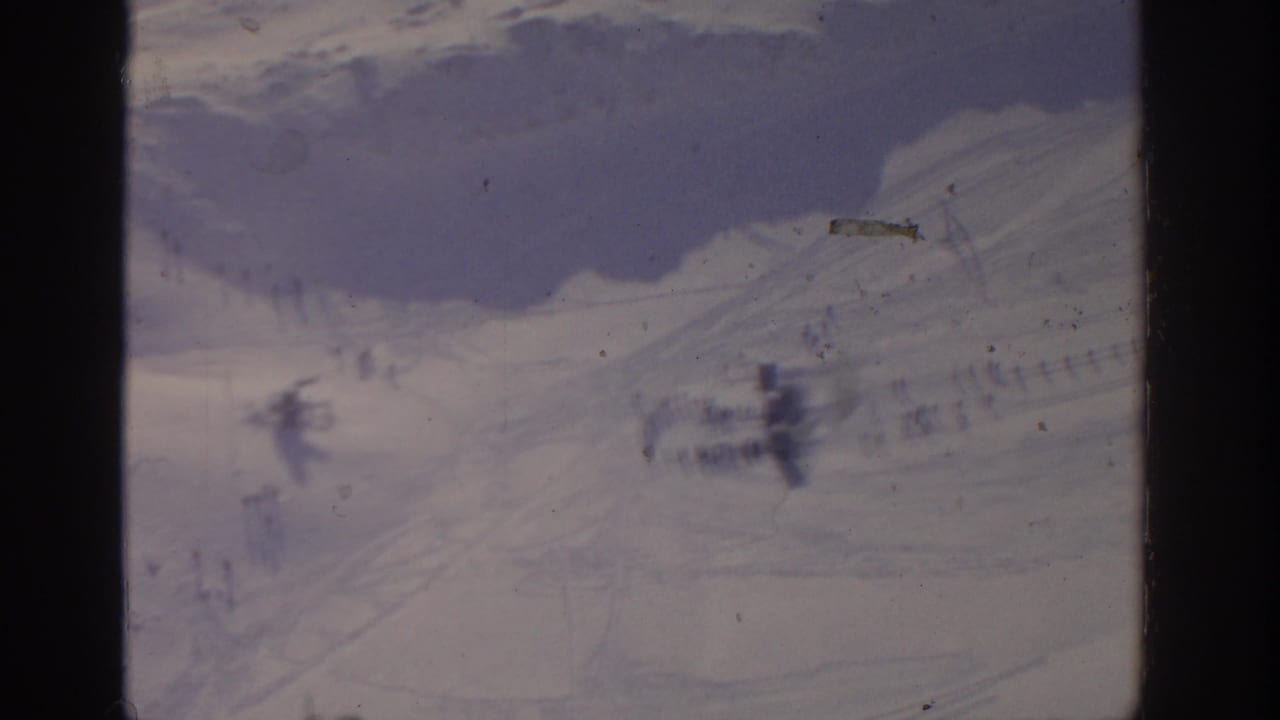 Distant snowy slope with small dark figures and faint tracks, seen through an 8mm frame.