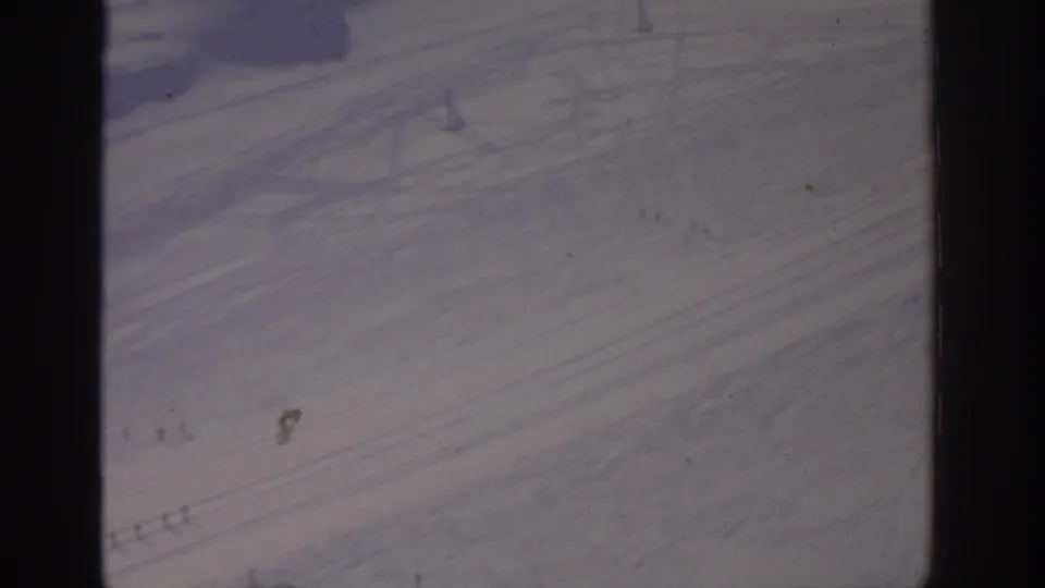 1963 archival 8mm frame, location suggested as Munich, showing a snowfield with diagonal tracks and small distant people on the slope.