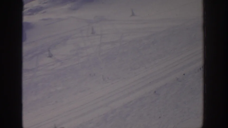 1963 archival 8mm frame showing a muted winter slope with scattered tracks and very small distant figures or objects in the snow.