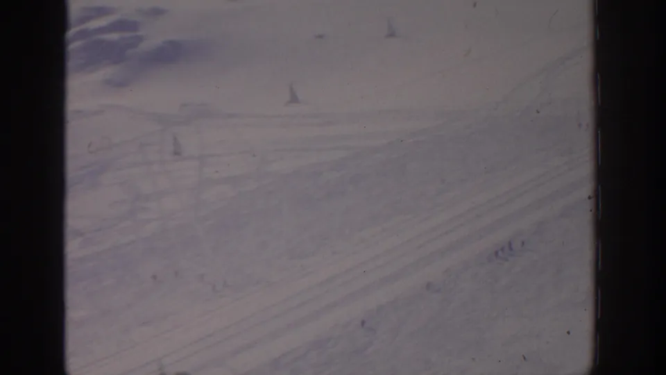 1963 archival 8mm frame, possibly Munich-area, showing curved snow tracks and tiny distant people across a pale winter landscape.