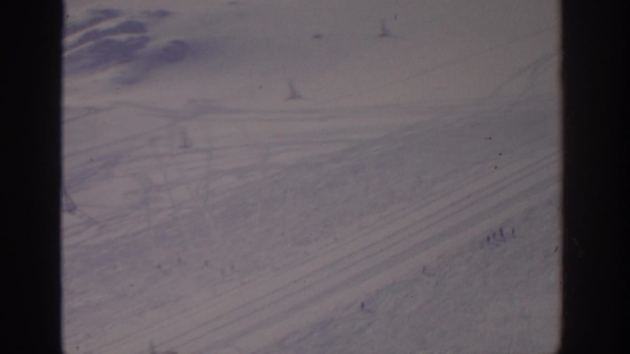 Snowy ski slope with tracks, lift towers, cable lines, and distant skiers.