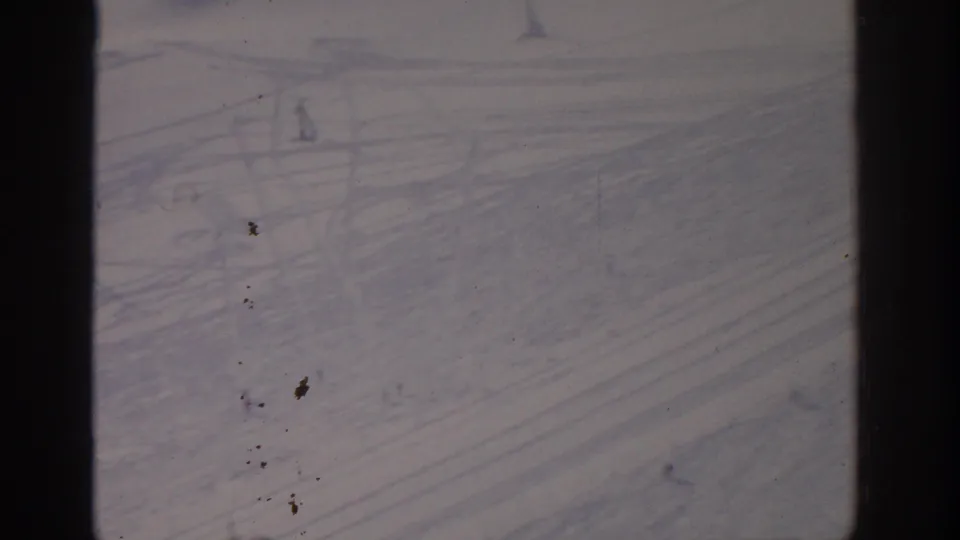 1963 8mm view near Munich of a tracked ski slope with distant skiers and lift supports partly softened by film exposure.