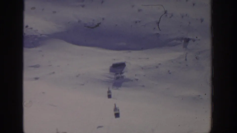 1963 home-movie frame near Munich showing two small aerial tram cabins over a snow-covered mountain landscape.