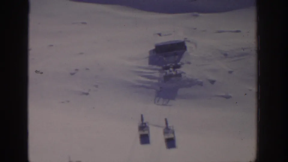 Closer 1963 Munich-area winter tram view with two cabins and a dark elevated structure set against a pale snowfield.
