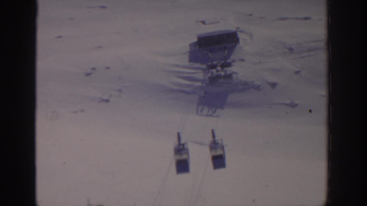 Two gondola cars hang below cables over a snowy mountainside near a lift station.