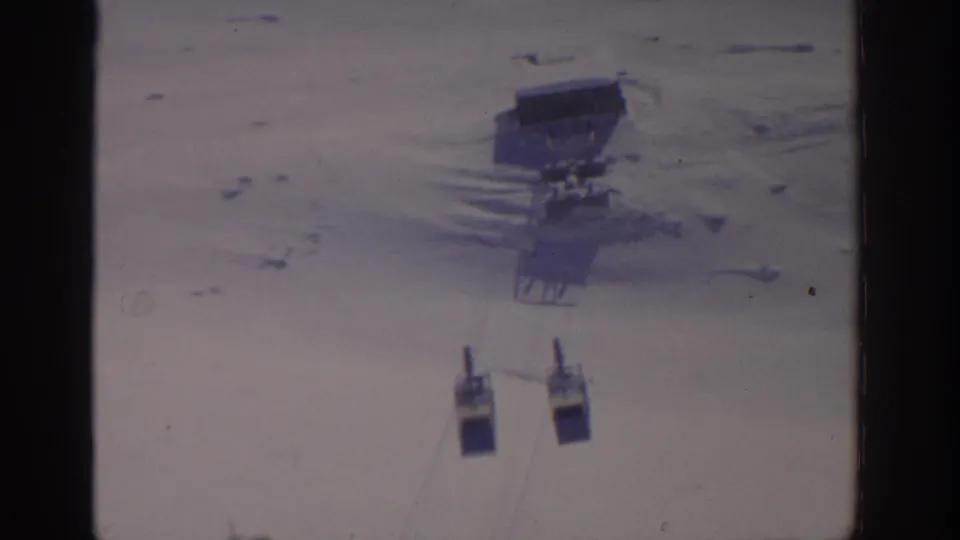 1963 8mm frame showing two gondola cars suspended over snow, with a lift station on the slope in the background.