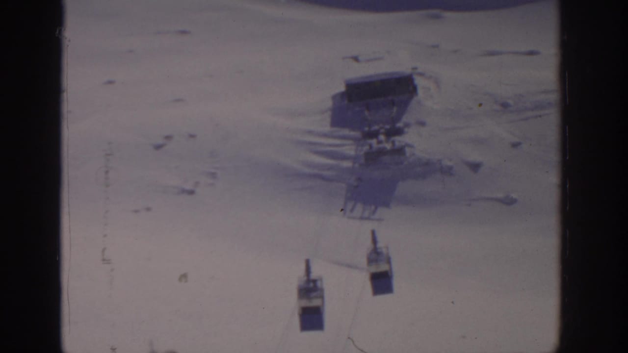 Gondola cars move along cables over snow with a station visible on the mountainside.