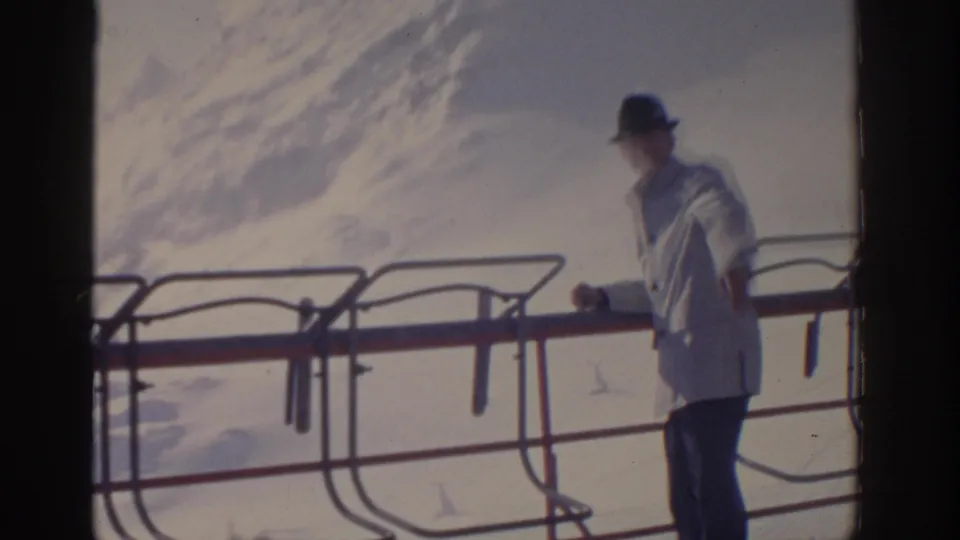 1963 8mm frame of an adult man in a light coat and dark hat standing at a railing against a snowy mountain backdrop.