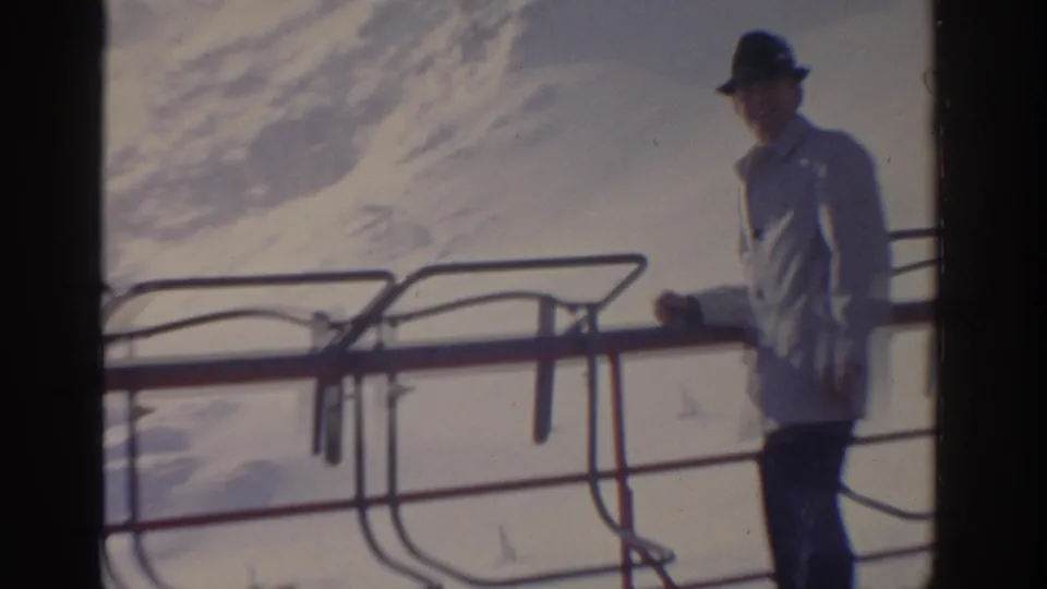 1963 home-movie frame showing an adult man in a light coat near a railing, with snowy mountain scenery behind him.