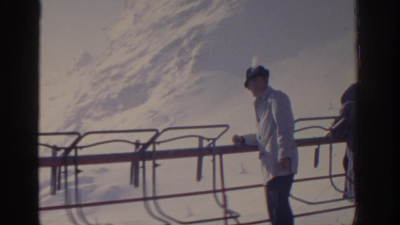 Man in a white coat and dark hat stands by ski-lift railing with snowy mountain behind him.