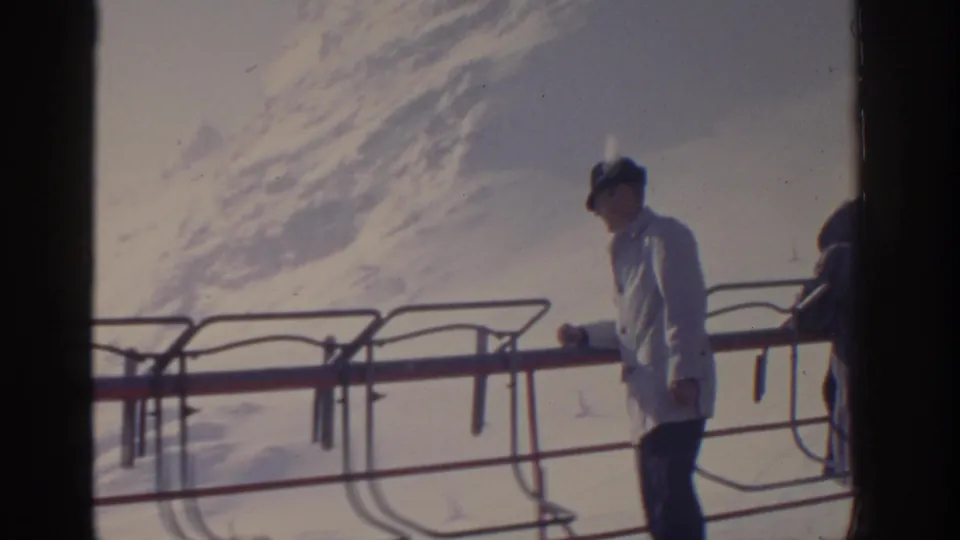 1963 Munich, Germany home-movie frame showing a man in a white coat near a metal ski-lift railing against bright snow.