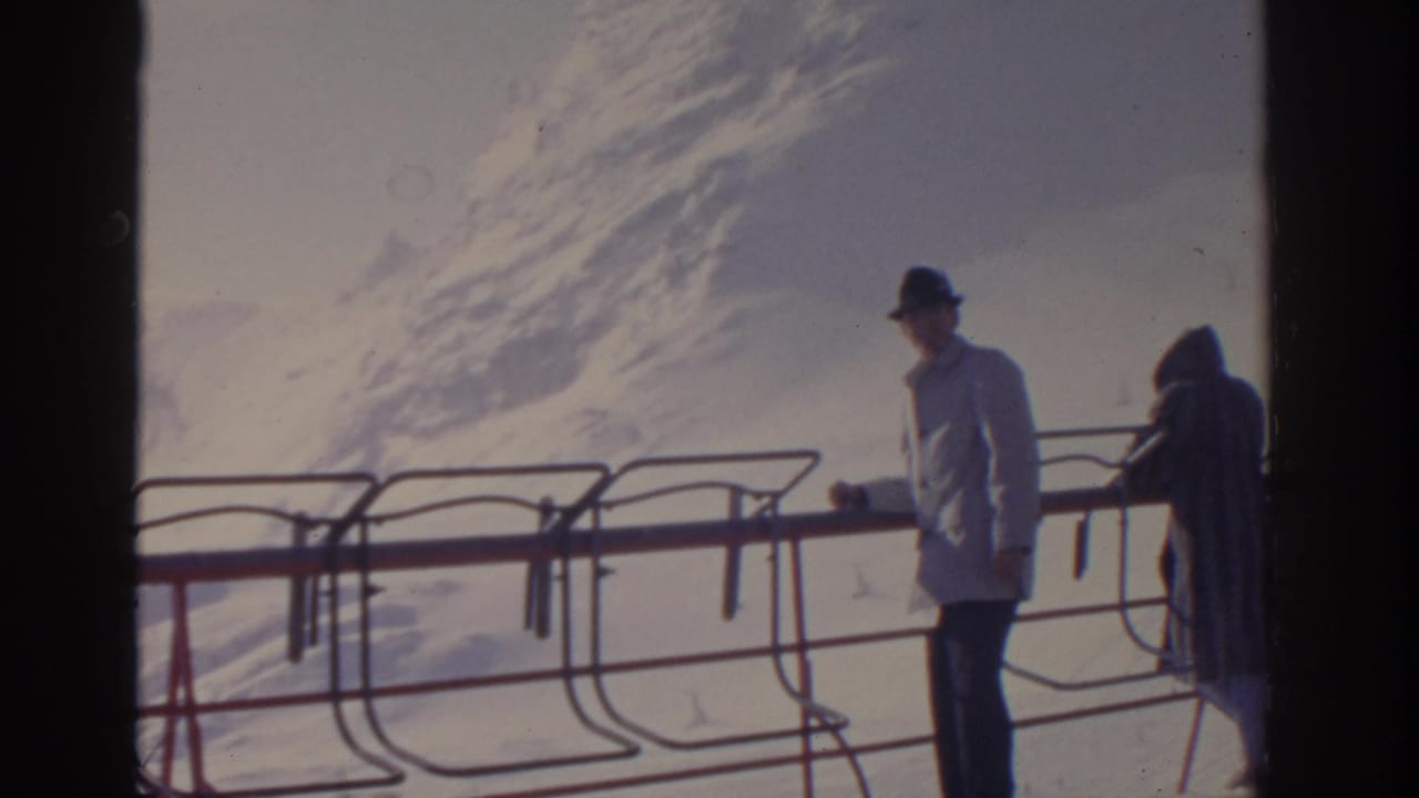 Man in a white coat stands near railing while a bundled person is partly visible at the right.