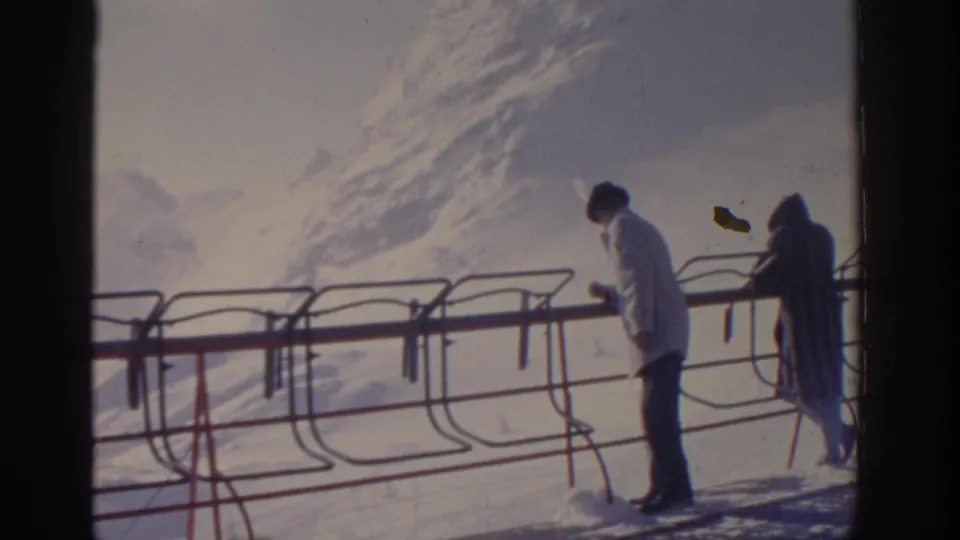 1963 Munich, Germany home-movie frame showing two people beside lift railings on a bright snowy platform.