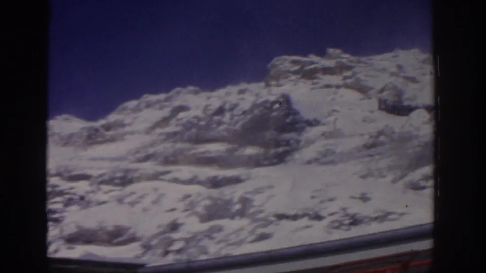 1963 8mm home-movie frame of a snow-covered mountain slope in Germany, with rocky outcrops and a deep blue sky.