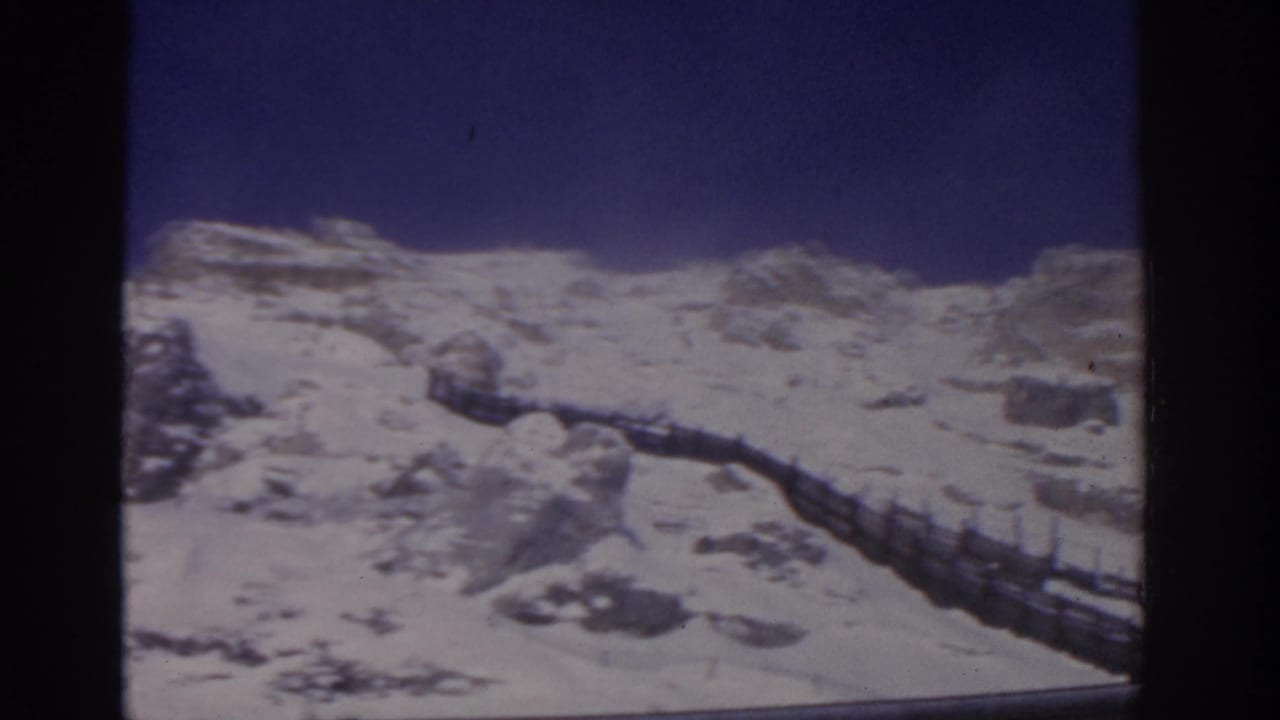 Snowy mountain terrain with a railing crossing the right side of the frame.