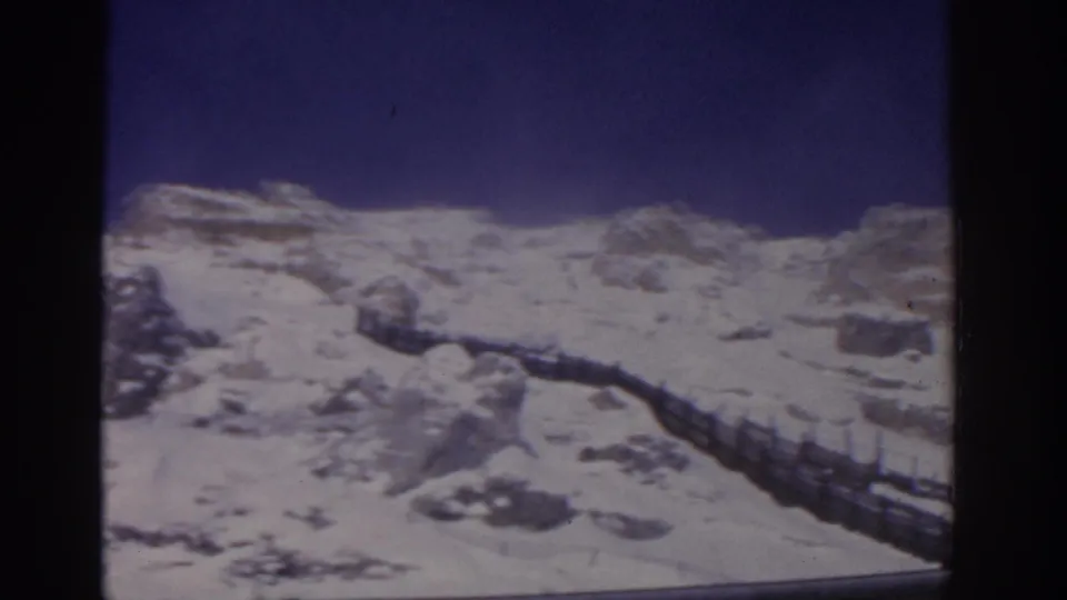 1963 8mm view of snowy mountain rocks and a railing or fence along the slope, with clear blue sky above.