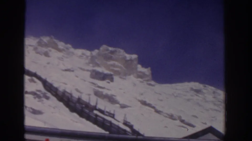 1963 8mm frame showing a railing on a snowy mountain slope, with rock formations rising against a clear sky.