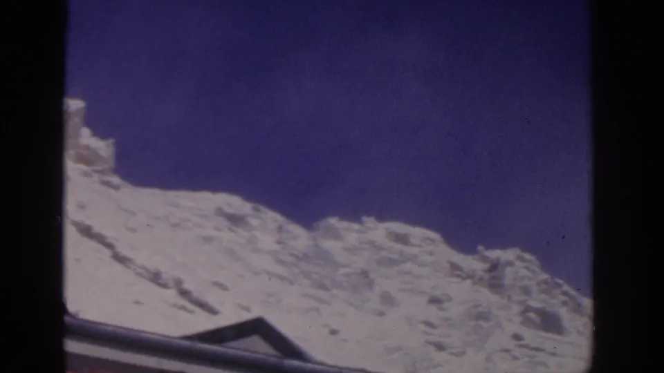 1963 8mm mountain view in Germany, showing a snow-covered rocky ridge and a partial roofline at the bottom edge.
