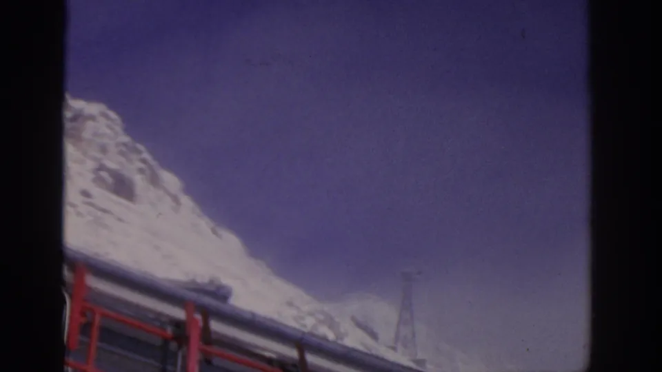 1963 8mm frame near Munich with a snowy alpine peak rising over a red-railed bus roof under a deep blue-purple sky.