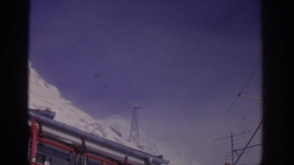 1963 8mm travel frame near Munich showing a snowy mountain edge, the bus roof, and a distant utility tower or post.