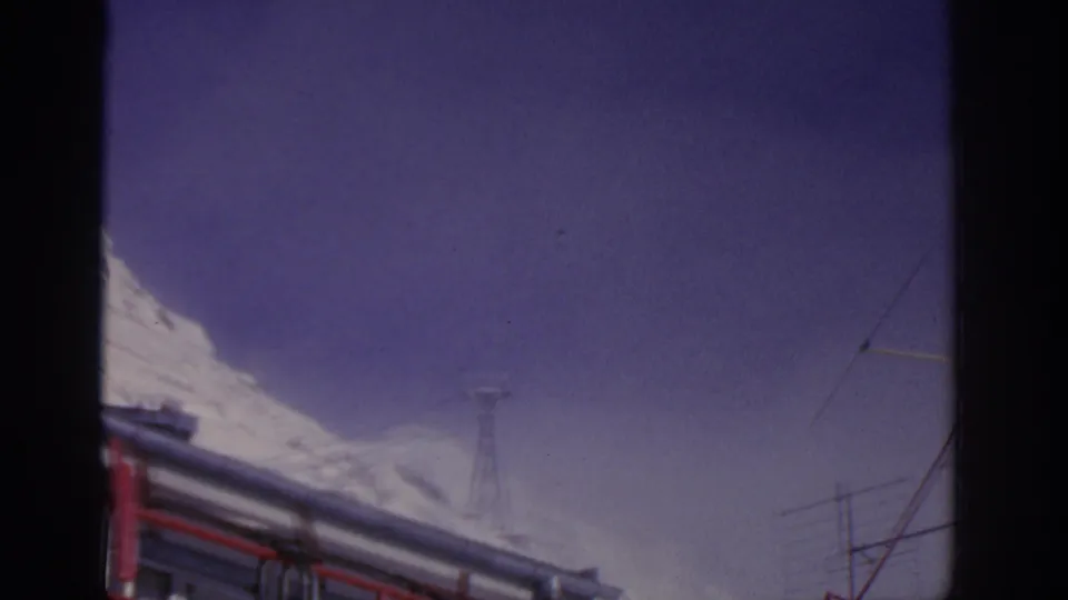 1963 8mm frame near Munich with bus roof details, snowy mountain edge, and distant tower or antenna structures against the sky.