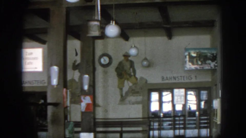 1963 Munich cafe or station-side interior with Bahnsteig 1 lettering, mural artwork, globe lamps, wall poster, and railings.