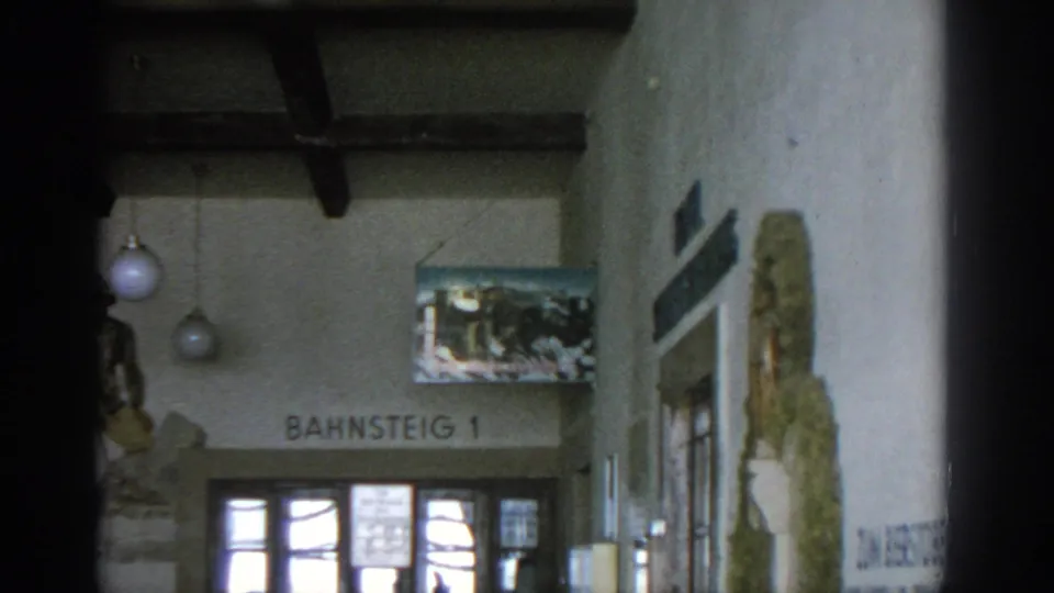 1963 Munich station interior panning closer to the Bahnsteig 1 sign, glass doors, globe lights, poster, and partial soldier mural.