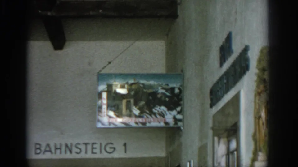 1963 Munich station detail showing the Bahnsteig 1 sign, overhead poster, glass doors, worn wall surface, and mural fragment at right.