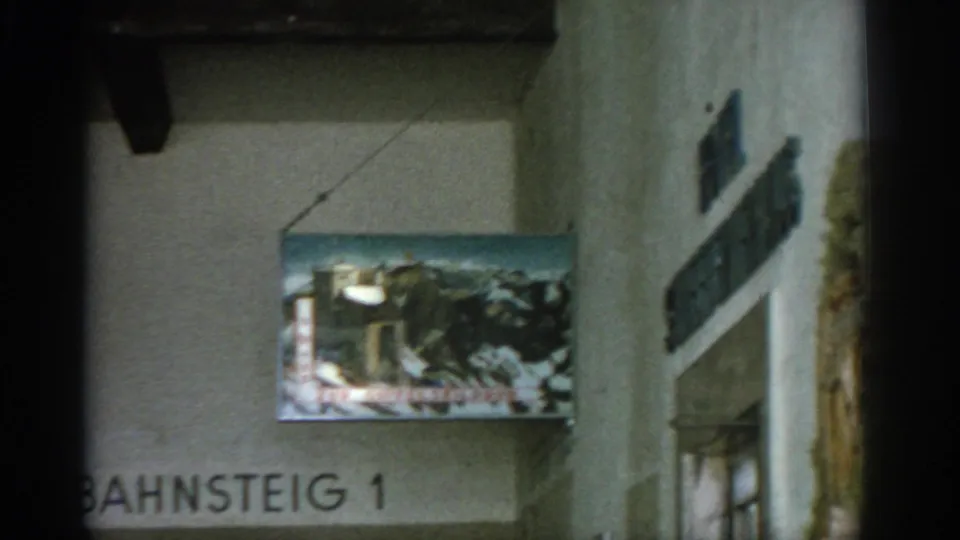 1963 8mm frame of German interior signage with a hanging mountain-scene sign and Bahnsteig 1 text on the wall.