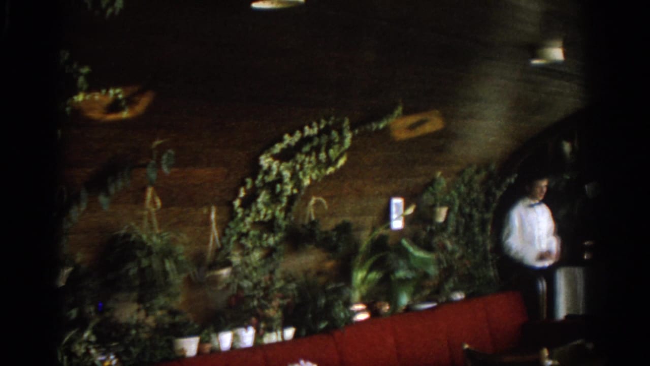 Dim ski lodge interior with waiter, red booth seating, plants, and warm lights.
