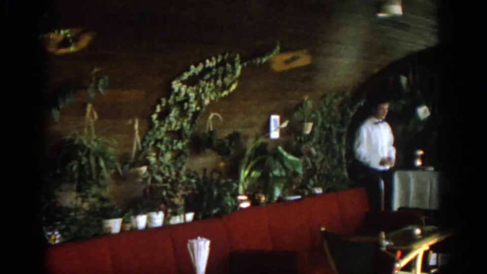 A waiter stands by a service table beside red booth seating and plants in a dim 1963 Munich resort interior.